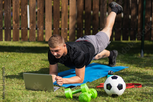 The young man goes in for sports in garden. Sportsman with blond hair does trains, makes swings and watch movie,watching a workout online on lawn