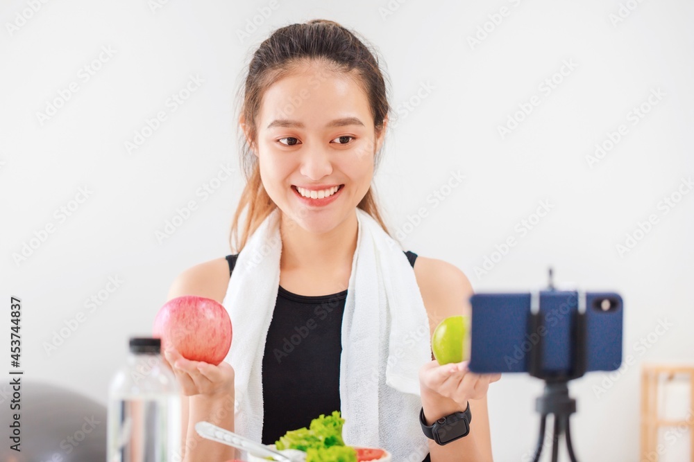 Beautiful asian woman healthy blogger is showing apple fruit and clean diet food. In front of the smartphone to recording vlog video live streaming at home.Fitness influencer on social media online. Beautiful asian woman healthy blogger is showing apple fruit and clean diet food. In front of the smartphone to recording vlog video live streaming at home.Fitness influencer on social media online.