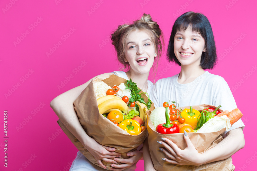 Two Positive Laughing Caucasian Girls Posing With Eco Paper Bags Filled With Grocery And Vegetables Over Pink Background.