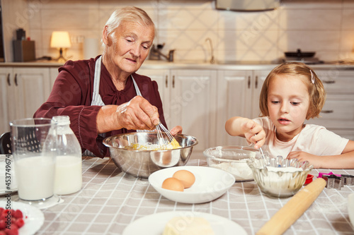 Fotografia Grandmother whipping eggs with flour while girl reaching for sugar