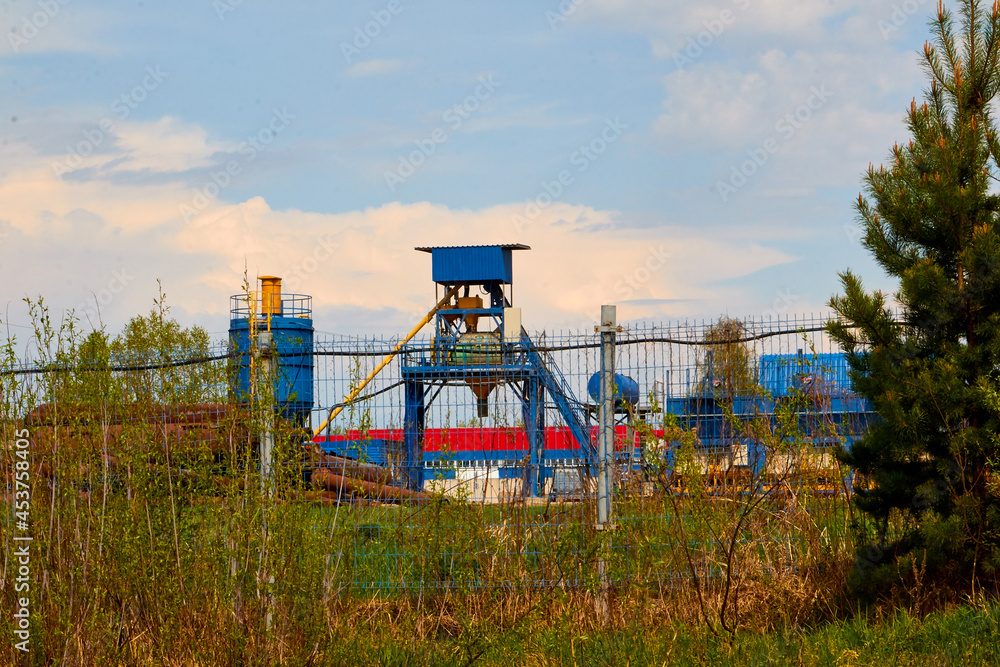Building of plant or factory in rural place framed by tree branches and shrubs and blue sky with white clouds Building of plant or factory in rural place framed by tree branches and shrubs and blue sky with white clouds
