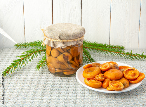 Marinated mushrooms orange milkcap or false saffron milkcap, Lactarius deterrimus in a glass jar and fresh picked mushrooms on plate, white wood board background. Side view. photo