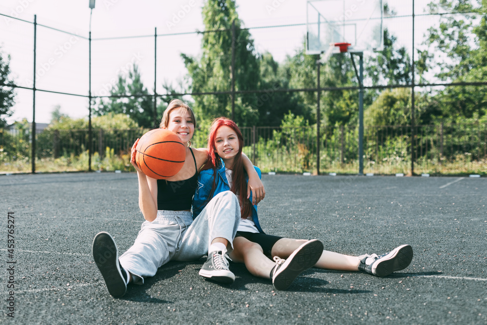 Two girls in sports clothes and with a basketball are chatting, sitting on the playground. Sports, competition, friendship Two girls in sports clothes and with a basketball are chatting, sitting on the playground. Sports, competition, friendship