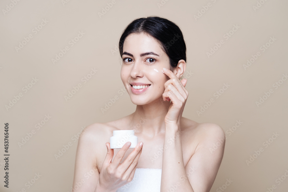 Close up beauty portrait of a laughing beautiful half naked woman applying face cream over beige background Close up beauty portrait of a laughing beautiful half naked woman applying face cream over beige background