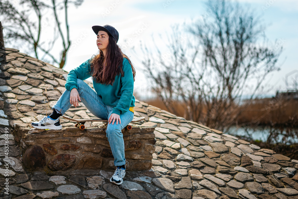 A young beautiful Caucasian woman sits on a skateboard and looks away. In the background, the blue sky with clouds and stone paving stones. Concept of sports lifestyle and street culture A young beautiful Caucasian woman sits on a skateboard and looks away. In the background, the blue sky with clouds and stone paving stones. Concept of sports lifestyle and street culture