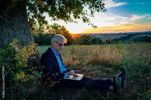 Senior businessman with laptop working under the magic tree at sunset
