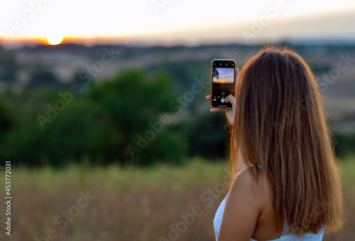 A young girl from behind with long blond hair, dressed in white, takes pictures of setting sun with her mobile phone