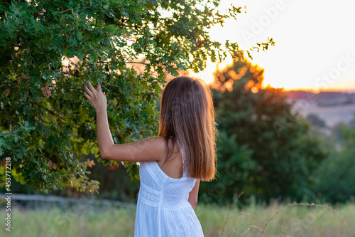 A young girl in profile with long blond hair, dressed in white, with her head turned to the setting sun, touches the leaves of the magic tree