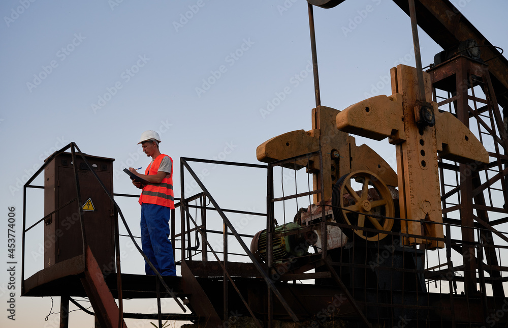 Oilman in work overalls, standing on oil rig and recording performance of pump. Oil and energy industry. Controller inspecting serviceability of pump rocker-machine. Oilman in work overalls, standing on oil rig and recording performance of pump. Oil and energy industry. Controller inspecting serviceability of pump rocker-machine.