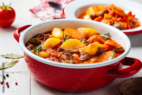 Beef stew with potatoes and carrots in tomato sauce in red pot, gray background.