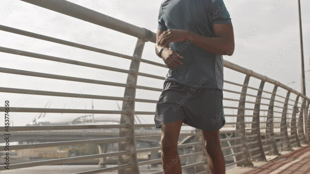 Midsection slowmo shot of fit African-American male athlete jogging along race track on sunny morning, looking at timer at his smartwatch Midsection slowmo shot of fit African-American male athlete jogging along race track on sunny morning, looking at timer at his smartwatch