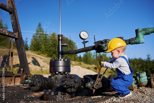 Fotografie Adorable little boy in construction helmet using industrial wrench while repairing pipe of oil well pump jack