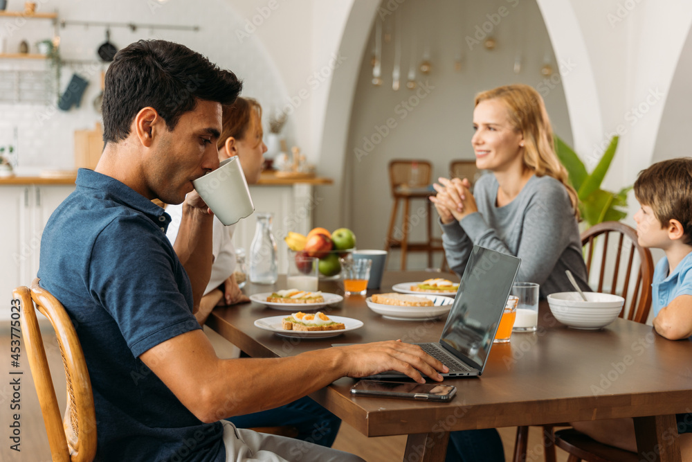 Handsome young man focusing and concentrating on working on laptop from home while eating breakfast with wife and cute little son Handsome young man focusing and concentrating on working on laptop from home while eating breakfast with wife and cute little son