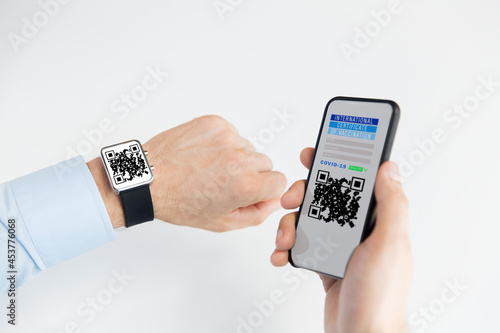technology and health care concept - man's hands holding smartphone with international certificate of vaccination and qr code on smart watch over white background