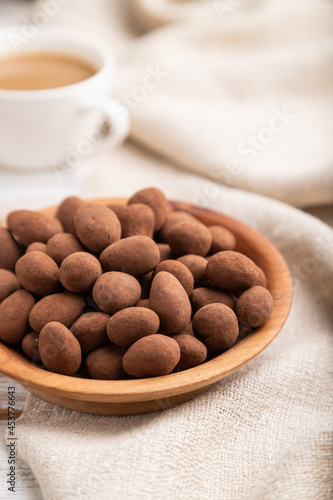Almond in chocolate dragees in wooden plate and a cup of coffee on white wooden background. Side view, selective focus.
