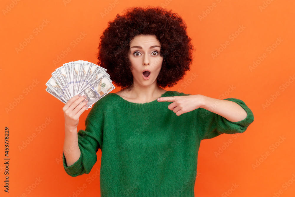 Portrait of shocked woman with curly hair wearing green casual style sweater holding fan of dollars and pointing at banknotes with open mouth. Indoor studio shot isolated on orange background.