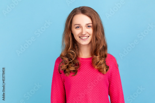 Portrait of smiling woman with wavy hair wearing pink pullover expressing positive emotions and happiness, looking at camera, being in good mood. Indoor studio shot isolated on blue background. © khosrork