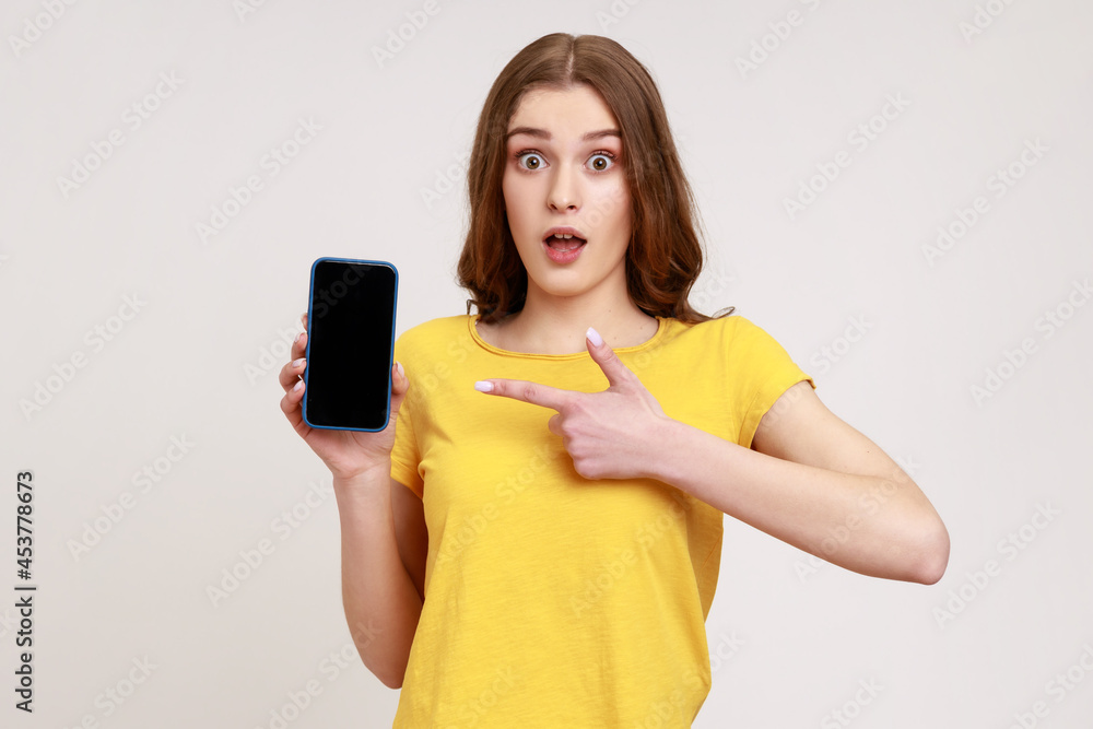 Portrait of shocked brown haired woman with wavy hair in yellow t-shirt pointing at cellphone and looking at camera with amazement and big eyes. Indoor studio shot isolated on gray background. Portrait of shocked brown haired woman with wavy hair in yellow t-shirt pointing at cellphone and looking at camera with amazement and big eyes. Indoor studio shot isolated on gray background.