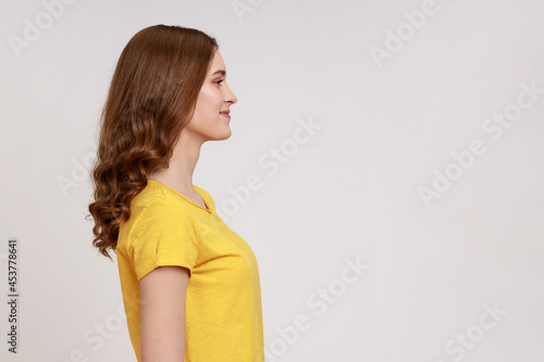 Side view of positive successful woman in yellow casual T-shirt standing with smile, looking happy and content, empty copy space for advertisement. Indoor studio shot isolated on gray background.