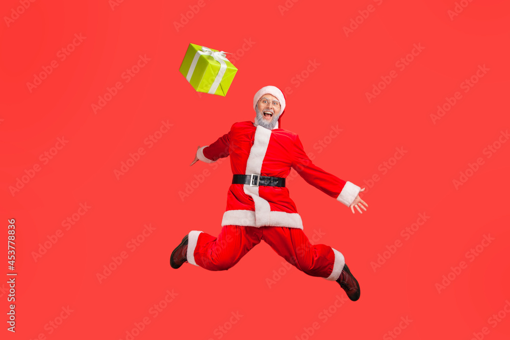 Full length of extremely happy elderly man with gray beard wearing santa claus costume jumping high and throwing gift up, spreading hands. Indoor studio shot isolated on red background.