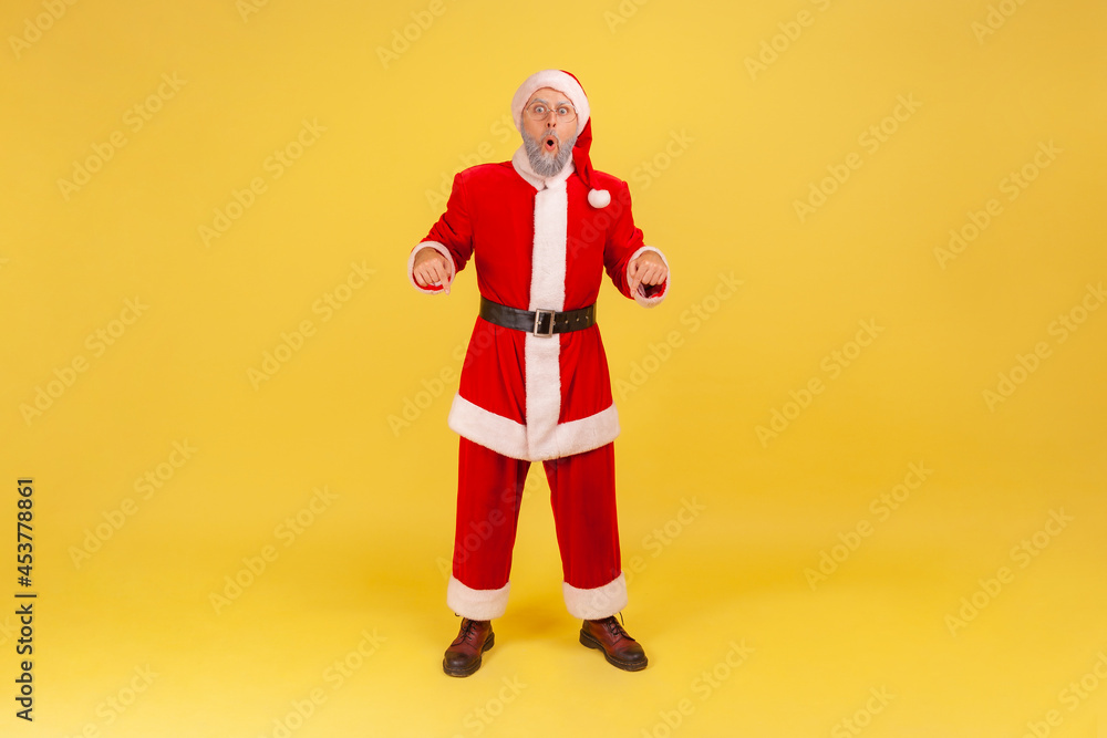 Full length portrait of elderly man with gray beard wearing santa claus costume standing with shocking expression and pointing down with both hands. Indoor studio shot isolated on yellow background. Full length portrait of elderly man with gray beard wearing santa claus costume standing with shocking expression and pointing down with both hands. Indoor studio shot isolated on yellow background.