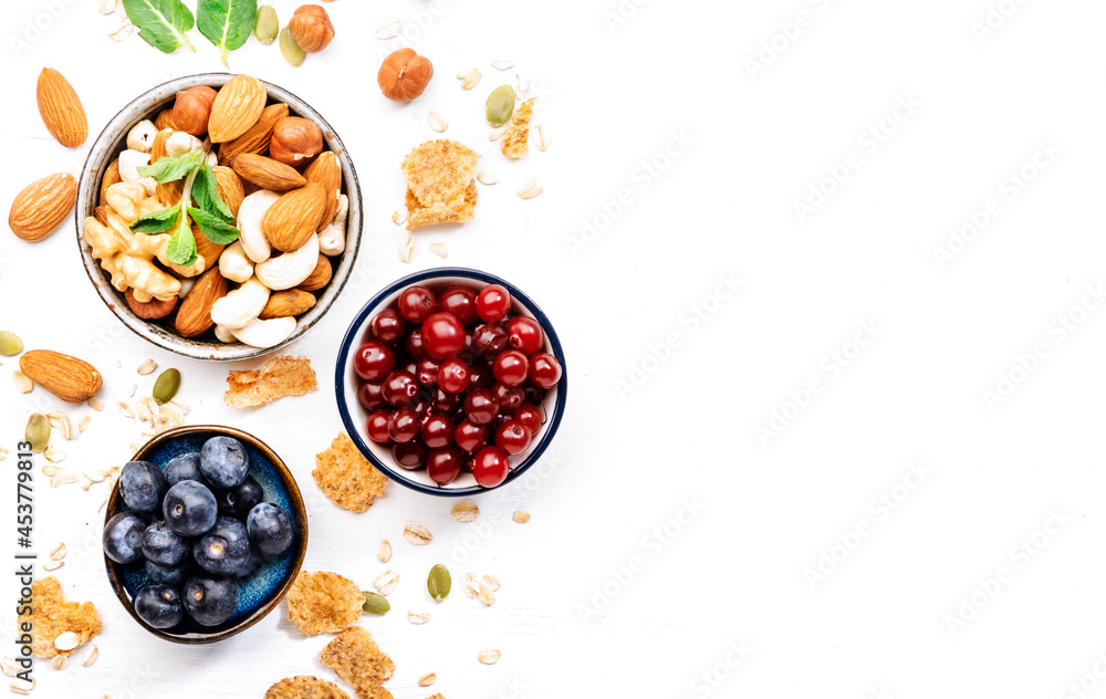 Muesli bowl and organic ingredients for healthy breakfast. Granola, nuts, blueberry, cranberry, oatmeal, greek yoghurt, whole grain flakes on white table Muesli bowl and organic ingredients for healthy breakfast. Granola, nuts, blueberry, cranberry, oatmeal, greek yoghurt, whole grain flakes on white table