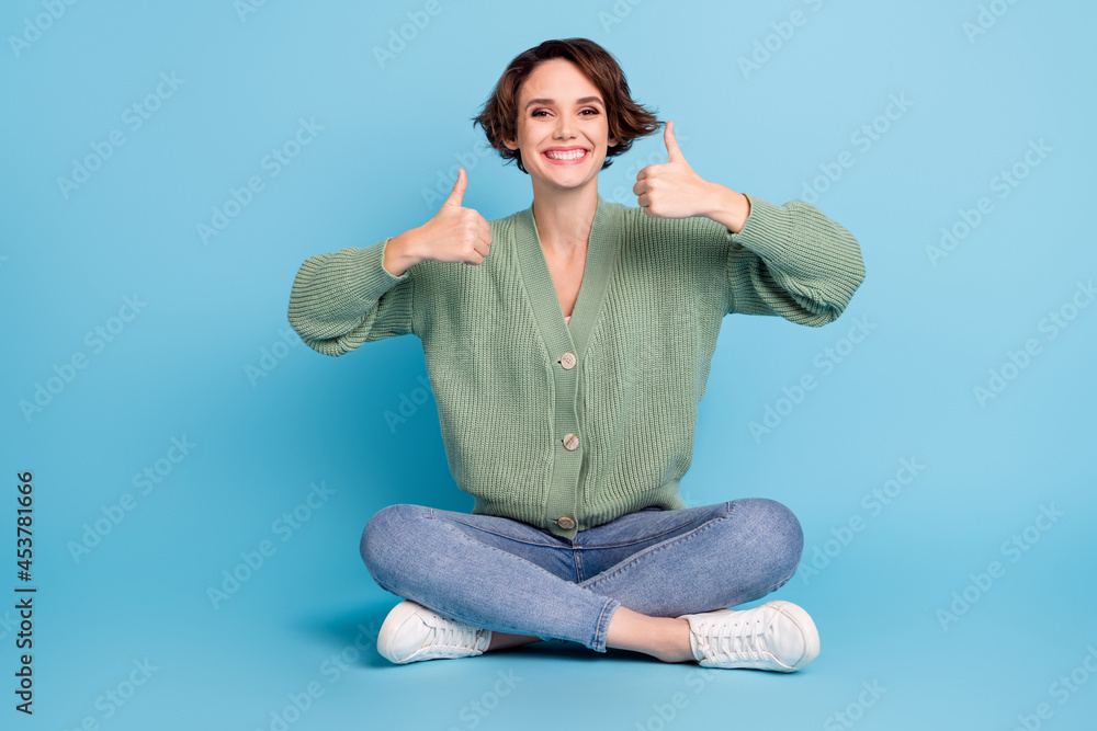Full length photo of shiny pretty young girl dressed green cardigan sitting floor showing thumbs up isolated blue color background Full length photo of shiny pretty young girl dressed green cardigan sitting floor showing thumbs up isolated blue color background