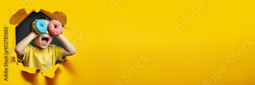 Happy cute boy is having fun played with donuts on Yellow background wall. Bright photo of a boy. Colored donuts