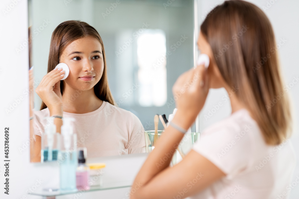 beauty, hygiene and people concept - teenage girl looking in mirror and cleaning face skin with cotton disc at bathroom beauty, hygiene and people concept - teenage girl looking in mirror and cleaning face skin with cotton disc at bathroom