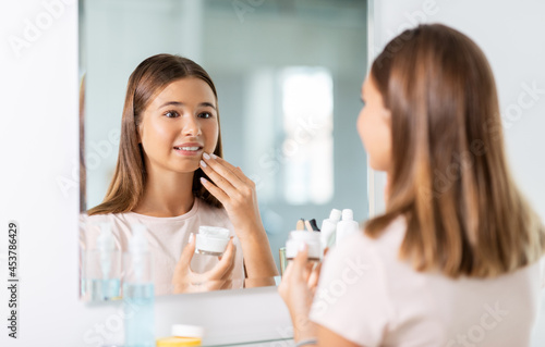 beauty, hygiene and people concept - teenage girl applying moisturizing cream looking in mirror at bathroom