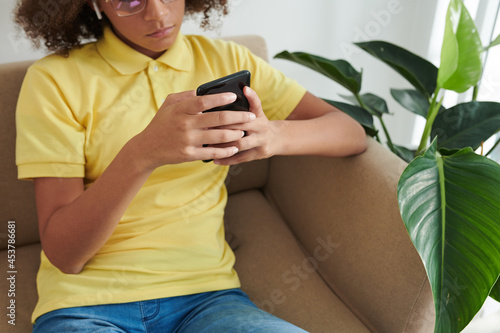 Serious teenage girl in glasses sitting on sofa in living room and answering text messages of friends