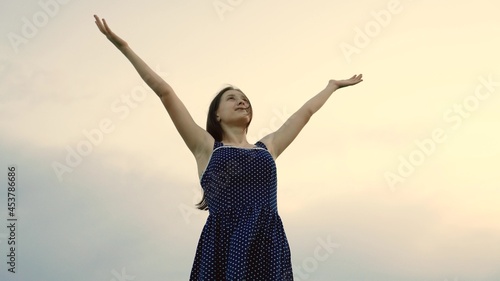 Young woman enjoying a calm evening in nature, breathing fresh air, in the summer park. People freedom concept. Free happy young girl tourist looking up with raised hands. Happiness under the sky.