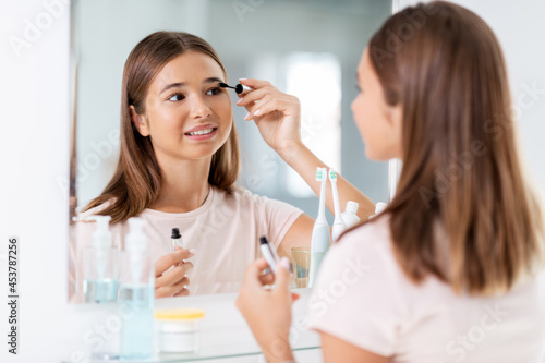 beauty, make up and cosmetics concept - teenage girl applying eye makeup with mascara and looking to mirror at home bathroom