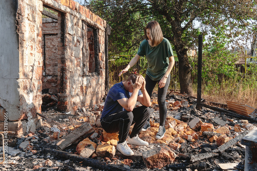 Young couple on the ruins of house destroyed with fire. Damaged window, walls and floor, burnt wooden frame. Man sits wrapping head in hands, woman calms him. Insurance or emergency concept.