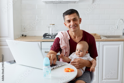 Happy smiling young adult father wearing maroon casual t shirt sitting at table in kitchen near notebook, holding infant in arms, looking at camera with positive expression.