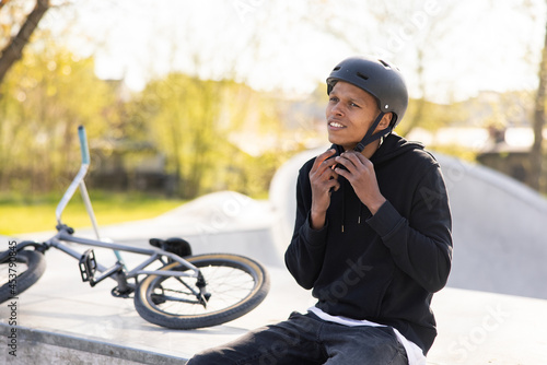 A cyclist who has fallen off bike, a bmx, and is sitting on a concrete ramp, gets ready to ride back on the track with friends, puts helmet on head and fastens it under his neck