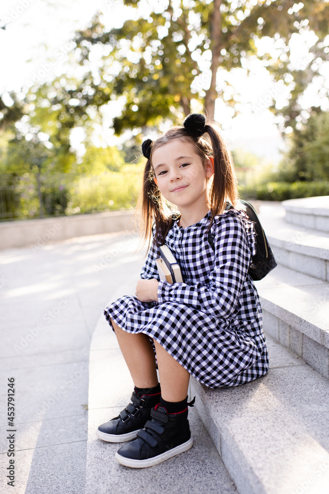 Cute young child girl 5-6 year old wear checkered black and white dress and backpack holding books sitting on stairs outdoors close up. Looking at camera. Back to school. Study concept.