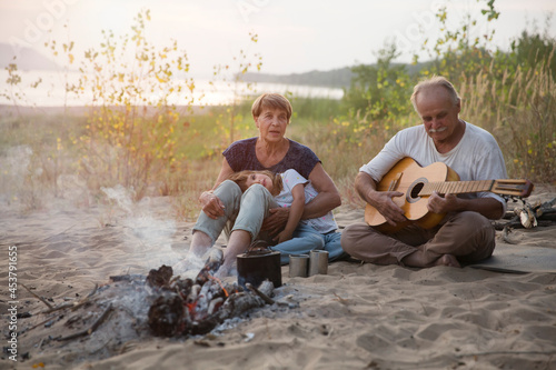 Grandparents and child granddaughter Camping holiday in the summer nature. Camping concept. senior manGrandfather playing guitar and singing song to his Family sitting on the beach near the fire
