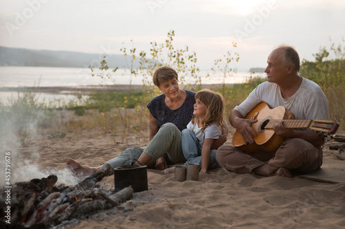 Grandparents and child granddaughter Camping holiday in the summer nature. Camping concept. senior manGrandfather playing guitar and singing song to his Family sitting on the beach near the fire  © ulza