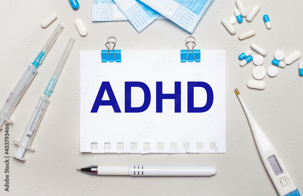 Fototapeta premium On a light gray background, blue medical masks, syringes, an electronic thermometer, pills, a pen and a notebook with the inscription ADHD. Medical concept