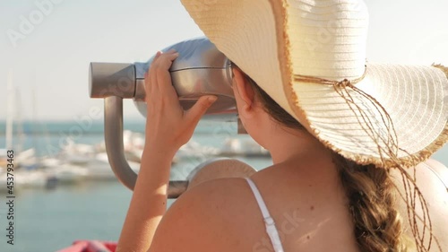 woman in beach clothes and hat looks through stationary binoculars. Binoscope on observation deck near the sea. seaport in Odessa. Expensive yachts on the pier. Commercial machine, vending business. photo