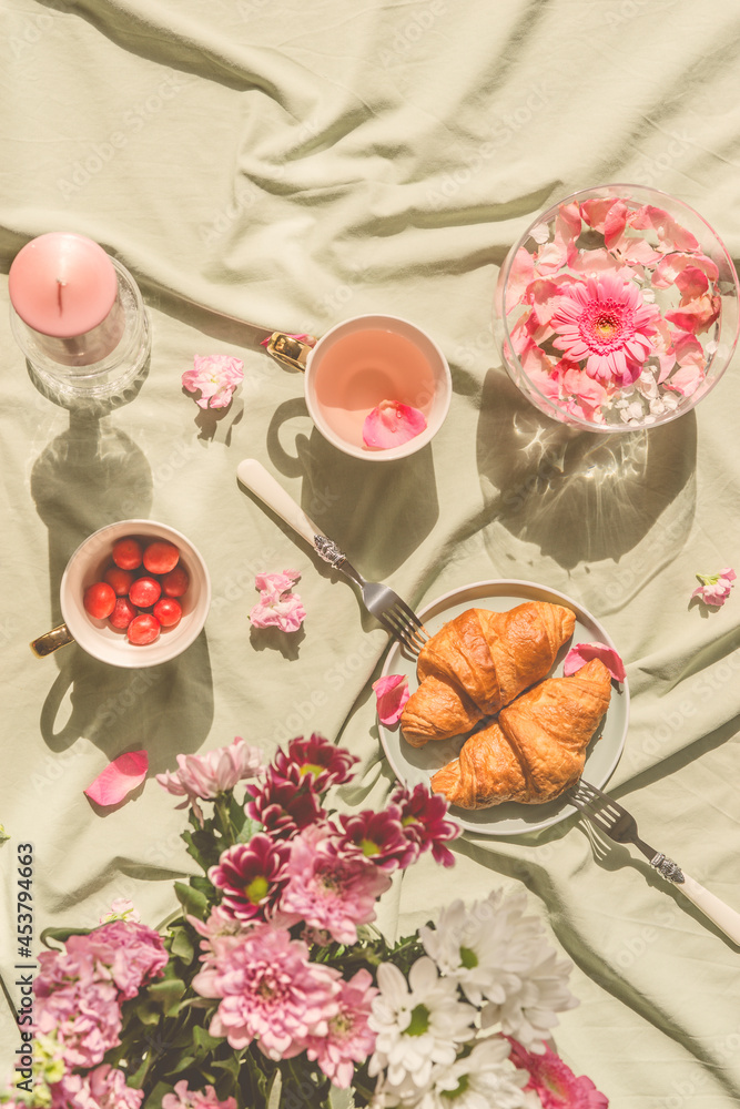 Sunny aesthetic picnic breakfast with flowers floating in crystal vases, croissants with tea from rose petals , bouquet of flowers and candles on blanket. Top view. Outdoor