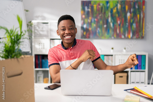Boy with dark skin in tshirt is satisfied with sold service products  happy with the monthly profits  sitting in front of computer victory dance  hands clenched into fists looking into camera  smiling