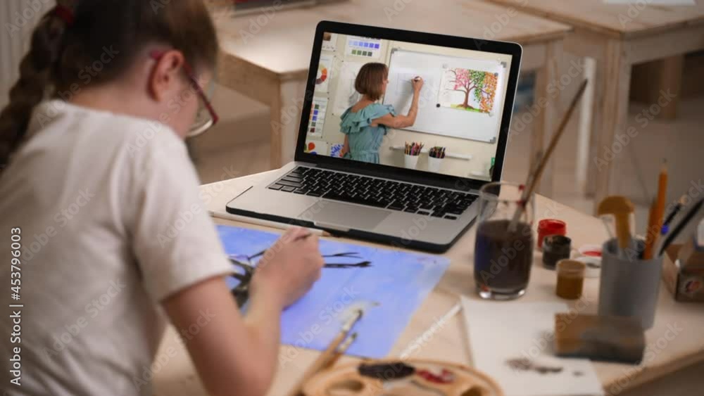 Girl drawing and studying remotely in front of laptop while sitting at table in home room spbas. Closeup view of schoolgirl draws in notebook and looks at computer screen, listens to woman teacher and Girl drawing and studying remotely in front of laptop while sitting at table in home room spbas. Closeup view of schoolgirl draws in notebook and looks at computer screen, listens to woman teacher and