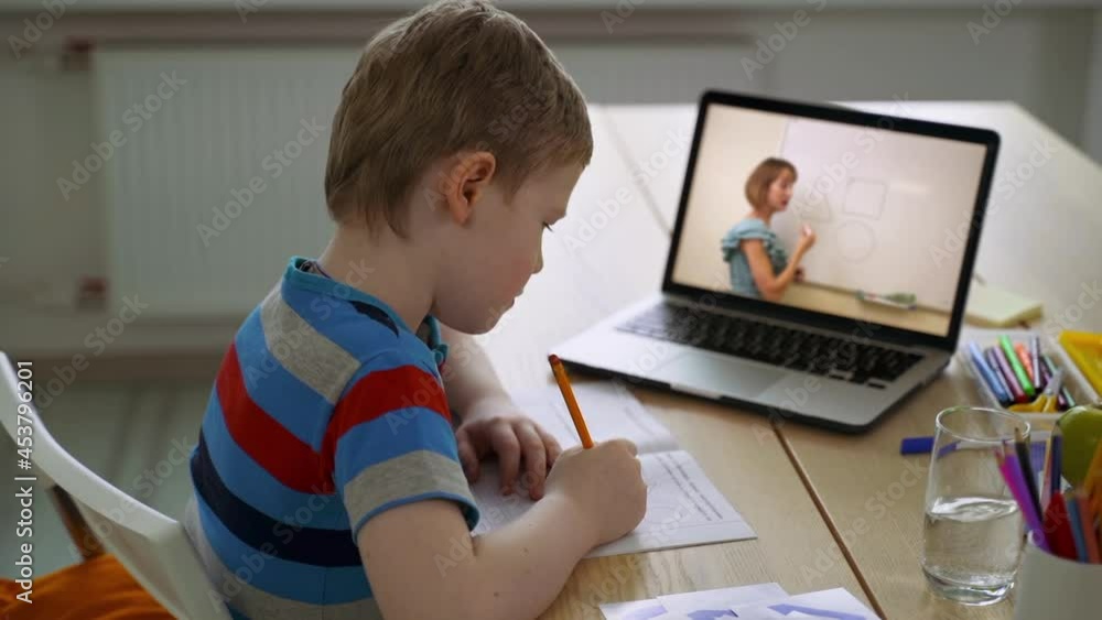 Schoolboy studying in online class and writing, sitting in front of laptop at home interior spbas. Close-up view of boy writes in notebook and sits at desk in room, looks at computer screen and Schoolboy studying in online class and writing, sitting in front of laptop at home interior spbas. Close-up view of boy writes in notebook and sits at desk in room, looks at computer screen and