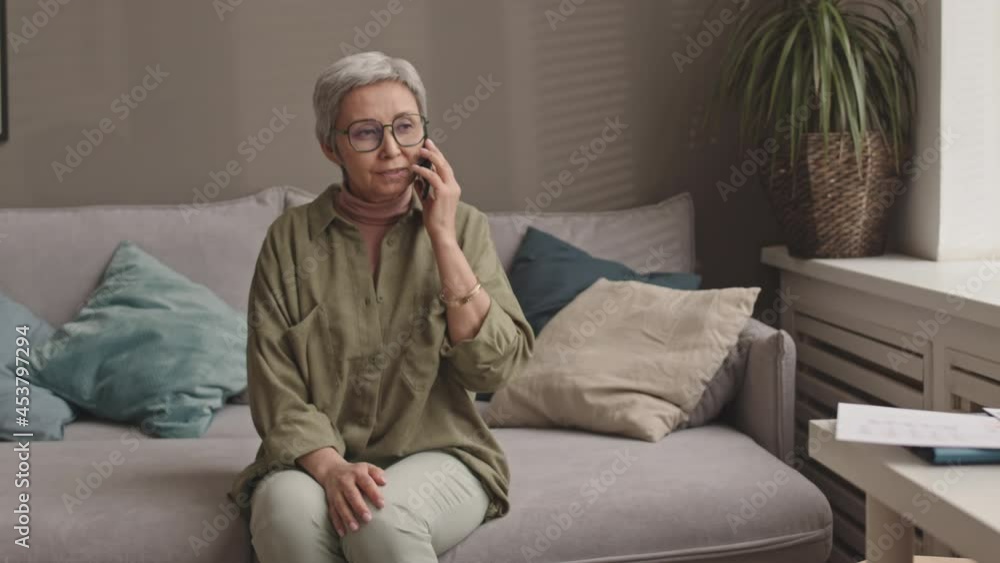 Medium long of successful older Asian woman sitting on couch of her home office, talking on mobile phone, then getting up and relocating to chair by desk and looking at documents Medium long of successful older Asian woman sitting on couch of her home office, talking on mobile phone, then getting up and relocating to chair by desk and looking at documents
