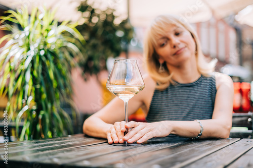 Happy adult mature woman sitting at table in bar outdoors with wine glasses and blurry restaurant background scene, drinking white wine. Summer sunny day on patio with plants. People lifestyle