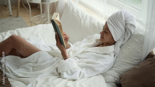 Beauty and body care at home. Spbd Happy African-American lady in bathrobe with towel turban on head reads book lying on comfortable bed near window