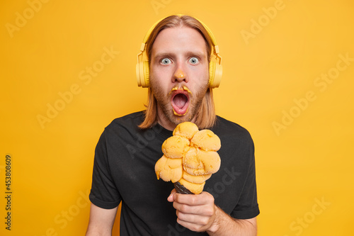 Emotional guy with red long hair stares shocked finds out horible news eats sweet summer dessert enjoys favorite songs from playlist isolated over yellow background. People and lifestyle concept photo