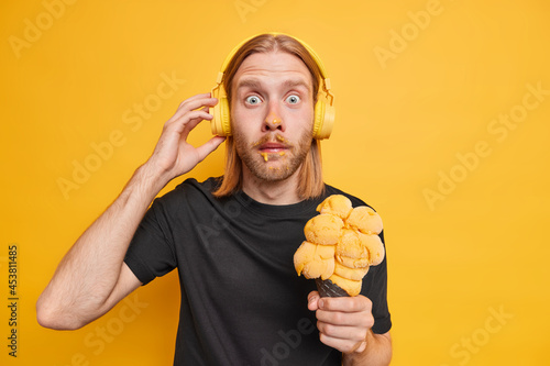 Surprised hipser guy smeared face as eats appetizing ice cream stares shocked spends summer time actively enjoys audio song wears casual black t shirt poses with favorite frozen dessert indoor photo